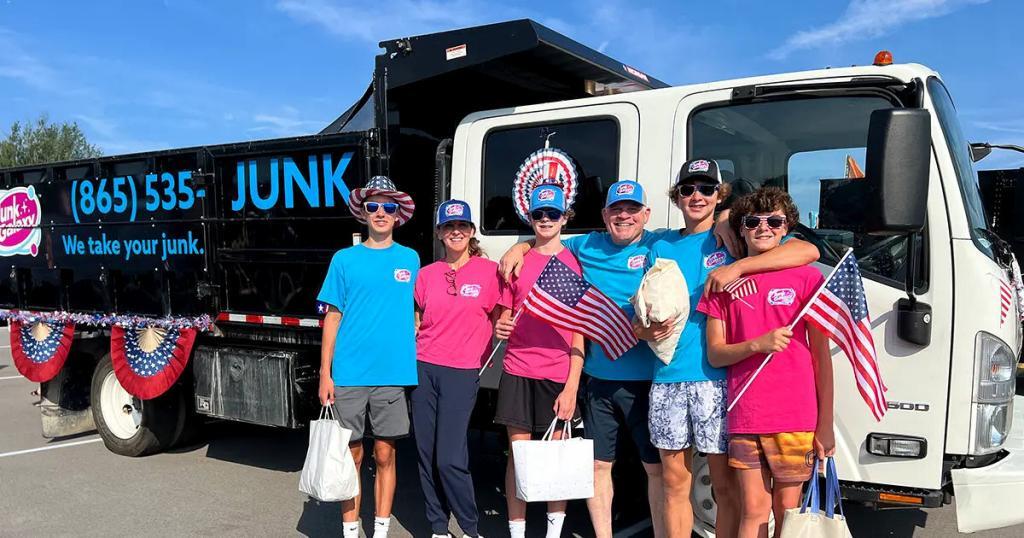 junk-galaxy-knoxville-a-group-of-people-in-vibrant-blue-and-pink-shirts-stand-beside-a-white-commercial-truck-with-junk-signage-holding-american-flags