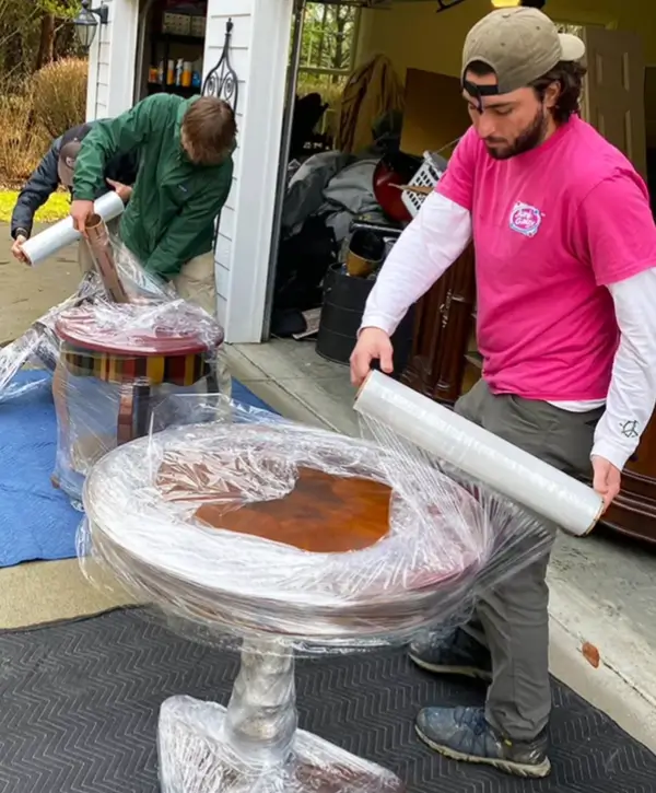 Workers wrapping wooden furniture for transport outside a garage.
