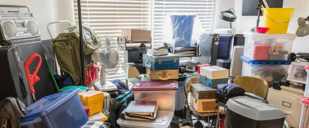 The interior of a hoarder house showing floor-to-ceiling piles of plastic bins, old radios, fans, and various household items blocking a window.