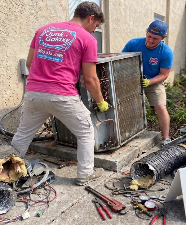 Two professionals lifting a rusty air conditioner off its concrete pad for AC disposal, with tools and detached ductwork scattered on the ground.